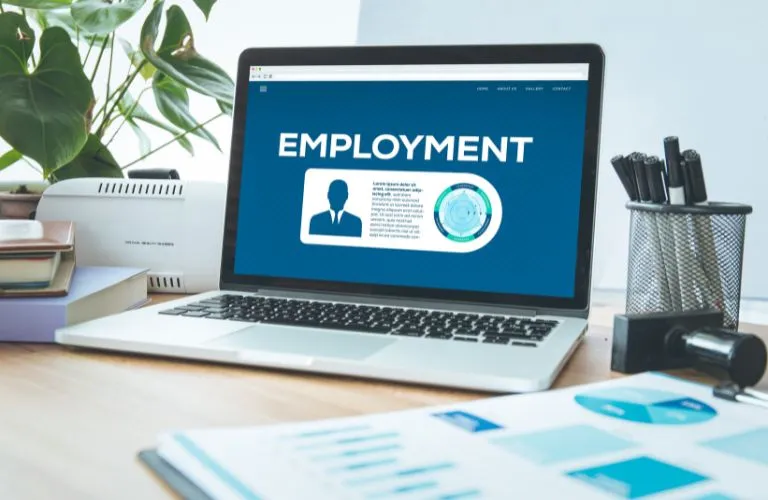 Laptop on wooden desk displaying an employment webpage, surrounded by office supplies and plants.