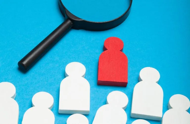 Black magnifying glass focusing on a red wooden figure among white wooden figures on blue background.