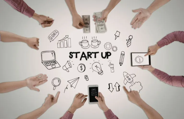 Overhead view of hands around startup-themed doodles, including money, laptop, and lightbulb icons.