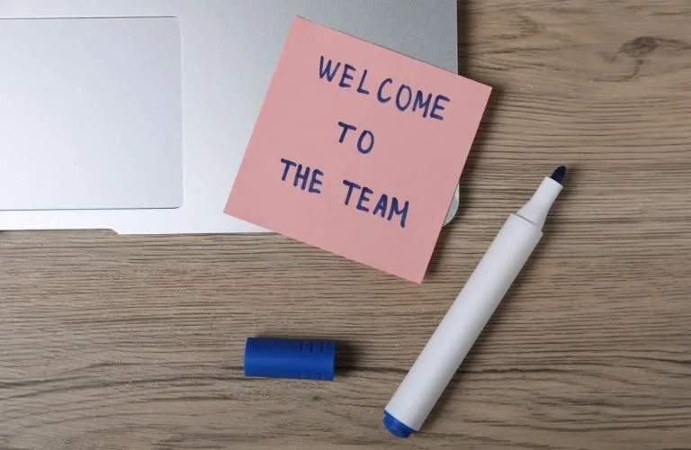 Pink sticky note with "Welcome to the team" text on a laptop keyboard, next to a blue marker.