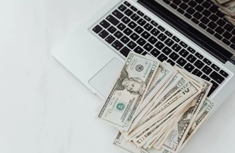 Stack of US dollar bills placed on a silver laptop keyboard on a white surface.