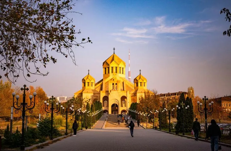 Sunlit Armenian church with golden domes, surrounded by trees and lampposts along a walkway.