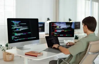 Person working on laptop and desktop computers displaying programming code in modern office.