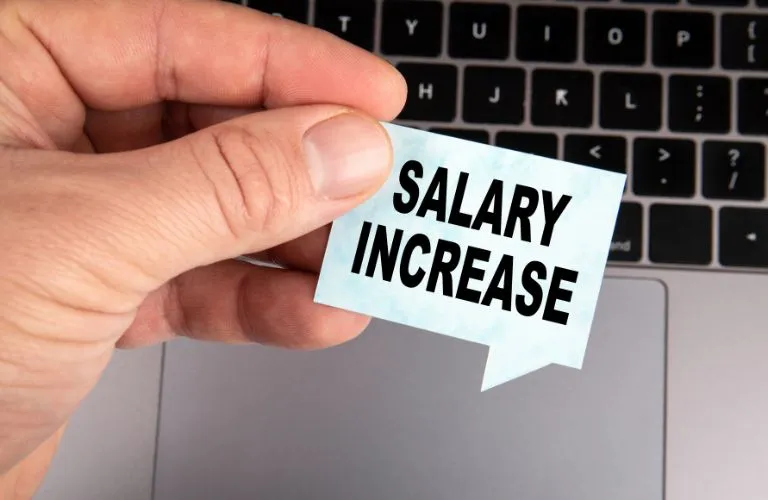 Hand holding a small blue speech bubble paper with bold black text "SALARY INCREASE" over a laptop keyboard background.