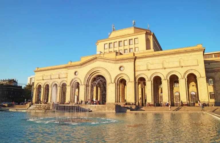 Large historic beige stone building with arches and a central fountain under clear blue sky.