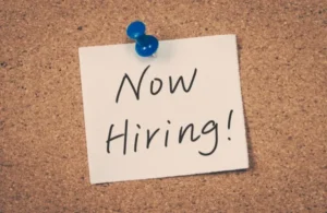 White note pinned with blue thumbtack on corkboard displaying "Now Hiring!" handwritten text.