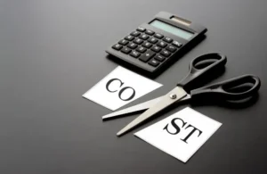 Black-handled scissors cutting a white paper with the word "COST" beside a black calculator on a dark surface.