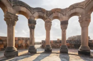 Stone arches and columns with intricate capitals under a cloudy sky at an ancient ruin site.