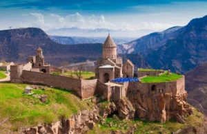 Ancient stone monastery with conical roof perched on cliff, surrounded by mountainous landscape.