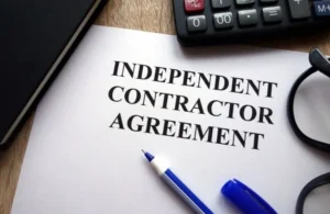 Document titled "Independent Contractor Agreement" on wooden desk with blue pen, calculator, and black eyeglasses.