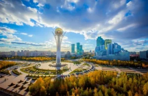Aerial view of a modern cityscape with a tall white tower topped by a golden sphere, surrounded by circular plazas, autumnal trees, and contemporary buildings under a partly cloudy blue sky.