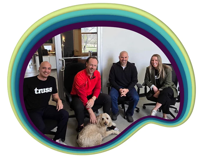 Four casually dressed adults seated indoors with a light-colored dog on the floor.