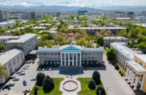 Aerial view of a neoclassical government building with white columns, surrounded by trees and cityscape, snow-capped mountains in background.