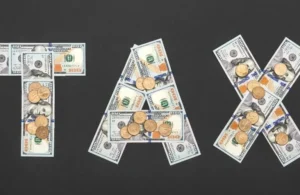 Letters "TAX" formed with US hundred-dollar bills and scattered coins on a dark background.