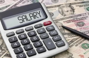 Close-up of a gray calculator displaying "SALARY" on the screen, placed on US dollar bills with a black pen nearby.