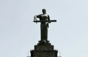 Statue of a woman holding a large sword, mounted on a stone pedestal against a clear sky.
