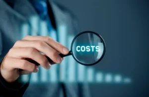Businessperson holding magnifying glass over the word "COSTS" with blurred bar chart background.